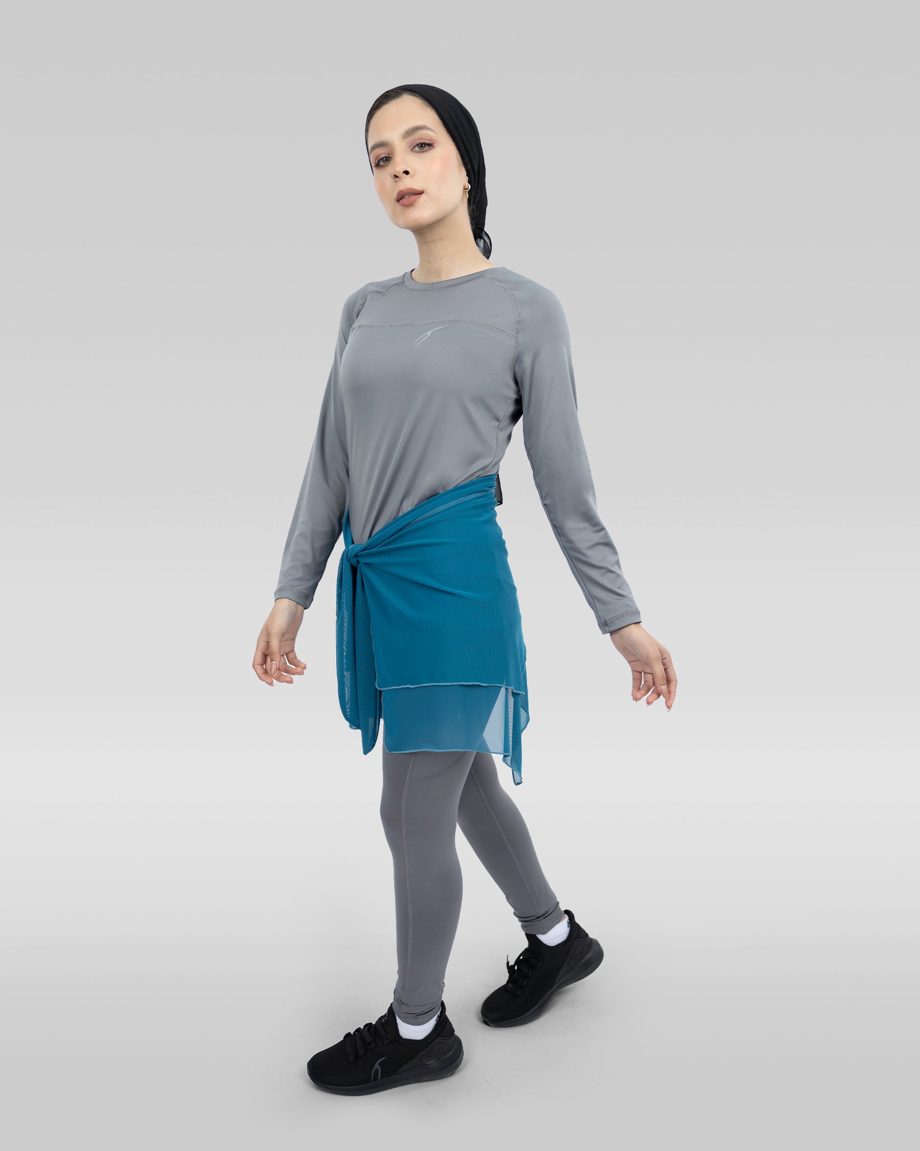 The image features a woman dressed in a gray and blue outfit, standing in a white room. She is wearing a gray shirt and a blue skirt, which is a combination of a dress and a skirt.