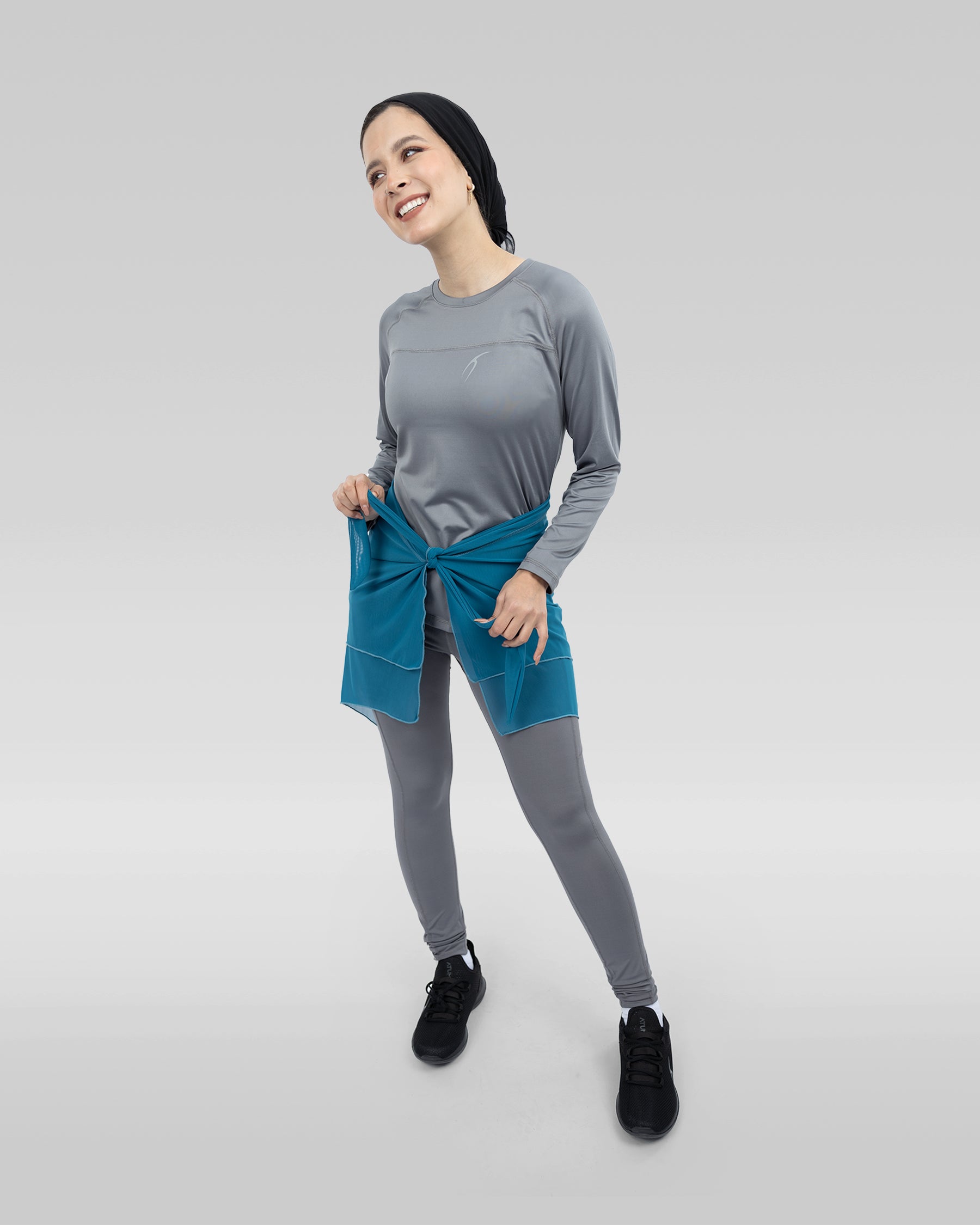 The image features a woman standing in a room, wearing a gray shirt and blue pants. She is posing for the camera, smiling and looking at the camera.